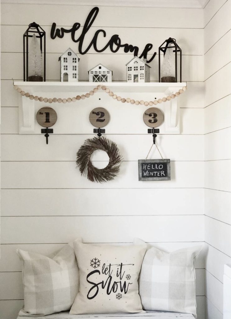 After photo demonstrating the transition from Christmas to Winter Decor. The space is transformed by removing red holiday accents and incorporating white and neutral colors, which infuse the area with a serene winter vibe. The decor includes a 'Welcome' sign above a shelf adorned with minimalist white houses and a simple wreath, complemented by white pillows with 'Let it Snow' script on the bench below, perfectly capturing the essence of winter simplicity.