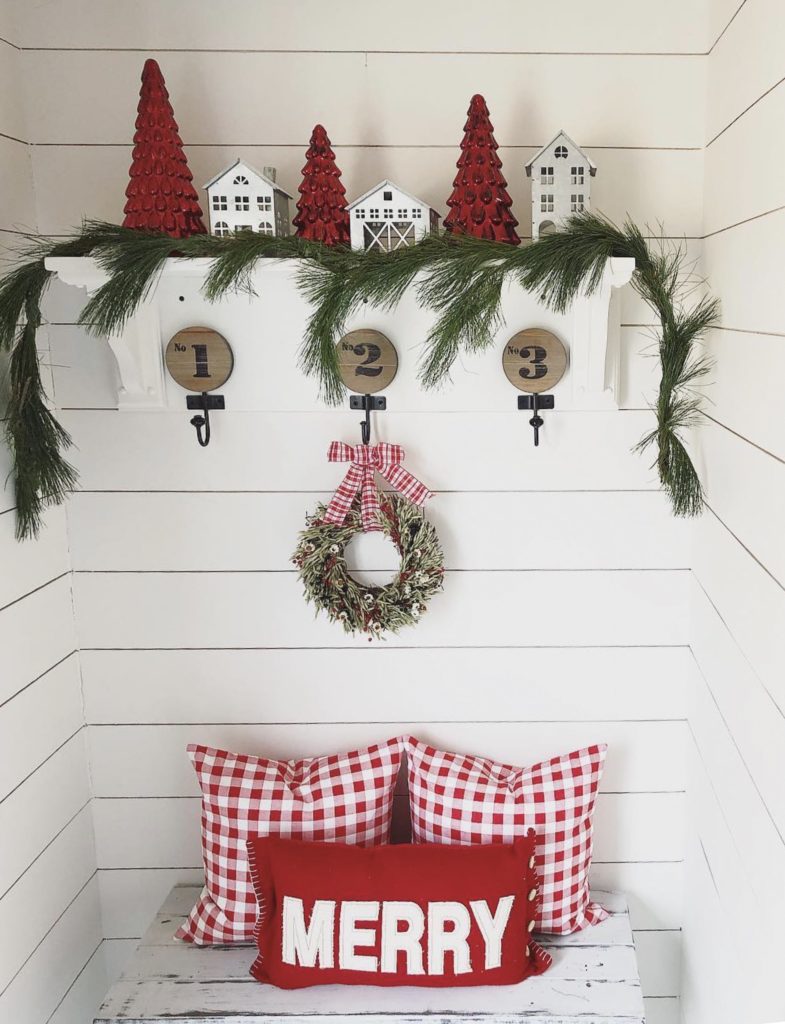 A festive Christmas display featuring a white bench with three built-in cubbies, each adorned with round wooden lids. Above the bench, a shelf holds three miniature white houses interspersed with red Christmas trees, all set against a backdrop of white shiplap walls. A lush green garland drapes over the shelf, accentuated by a small wreath with red and white decorations hanging in the center. On the bench, two red and white gingham pillows flank a larger red pillow with the word 'MERRY' in white lettering, creating a cozy and inviting holiday scene