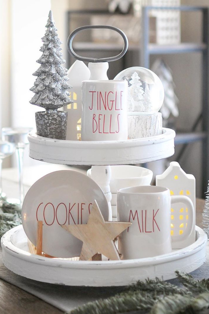 Two tiered white wooden tray styled with white Rae Dunn Christmas dishes and other Christmas touches, like a white snow globe. Via dogsndecor.com