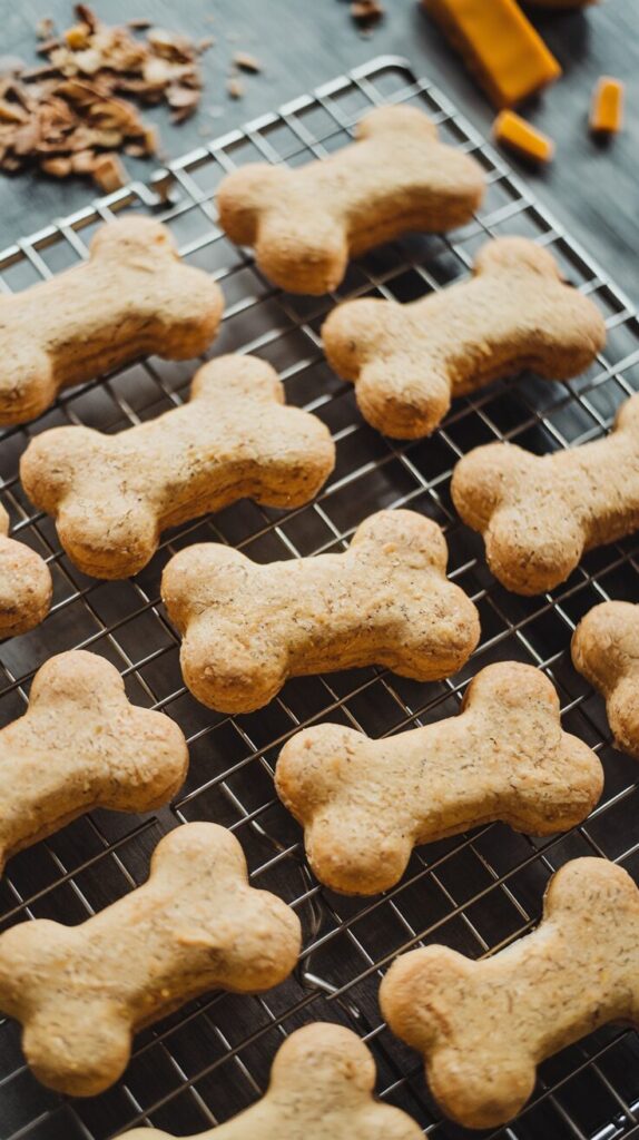 A picture of freshly baked, bone-shaped cheesy dog biscuits cooling on a wire rack. The biscuits are perfectly golden with a slightly crispy texture. Surrounding the rack are scattered ingredients like shredded cheddar cheese and whole wheat flour, adding context to the baking process. 